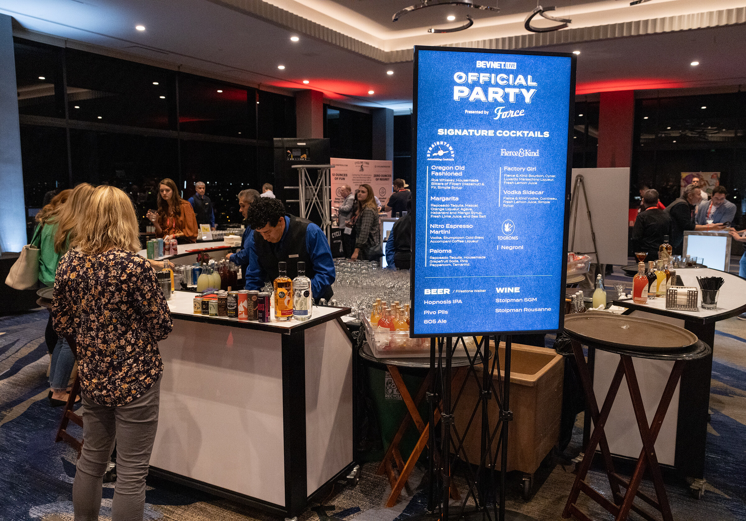 A bar with a menu screen reading Official Party Signature Cocktails and a bartender preparing drinks.