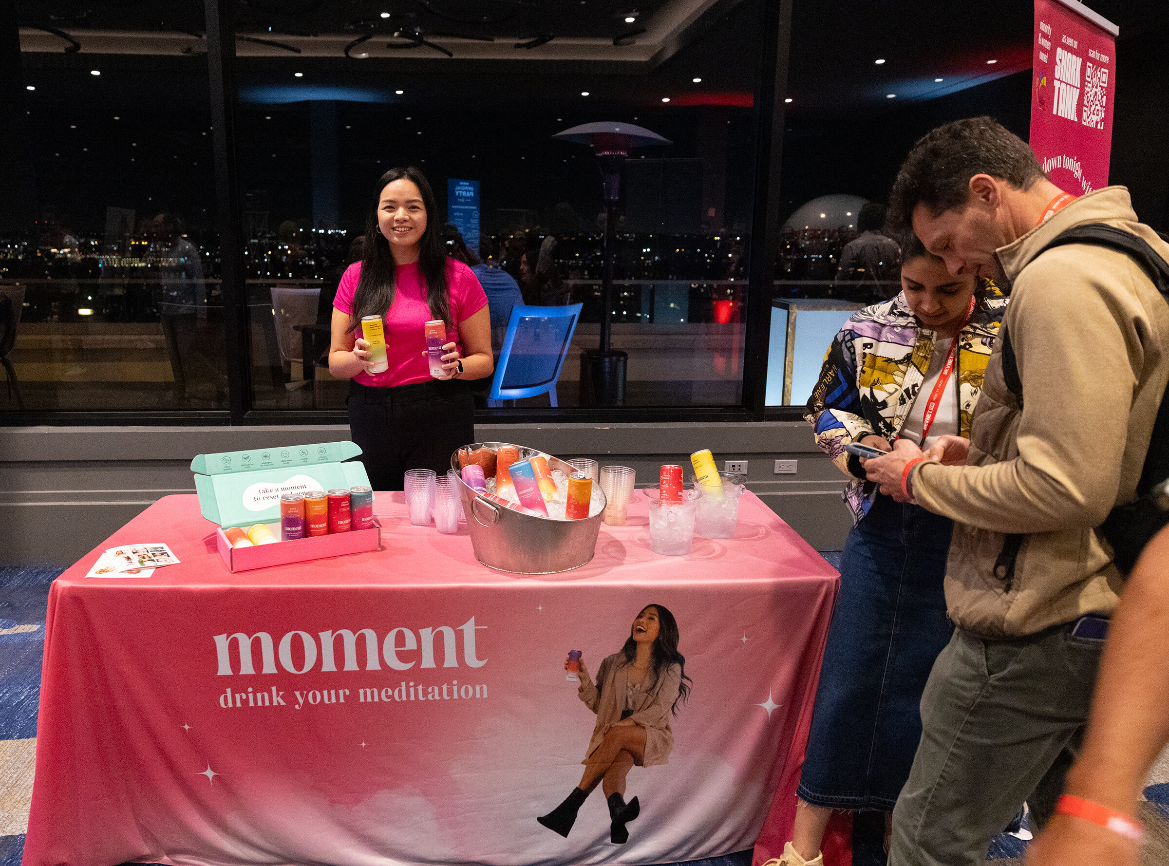 A woman in a pink top stands behind a pink table that reads moment, with attendees in front of her.