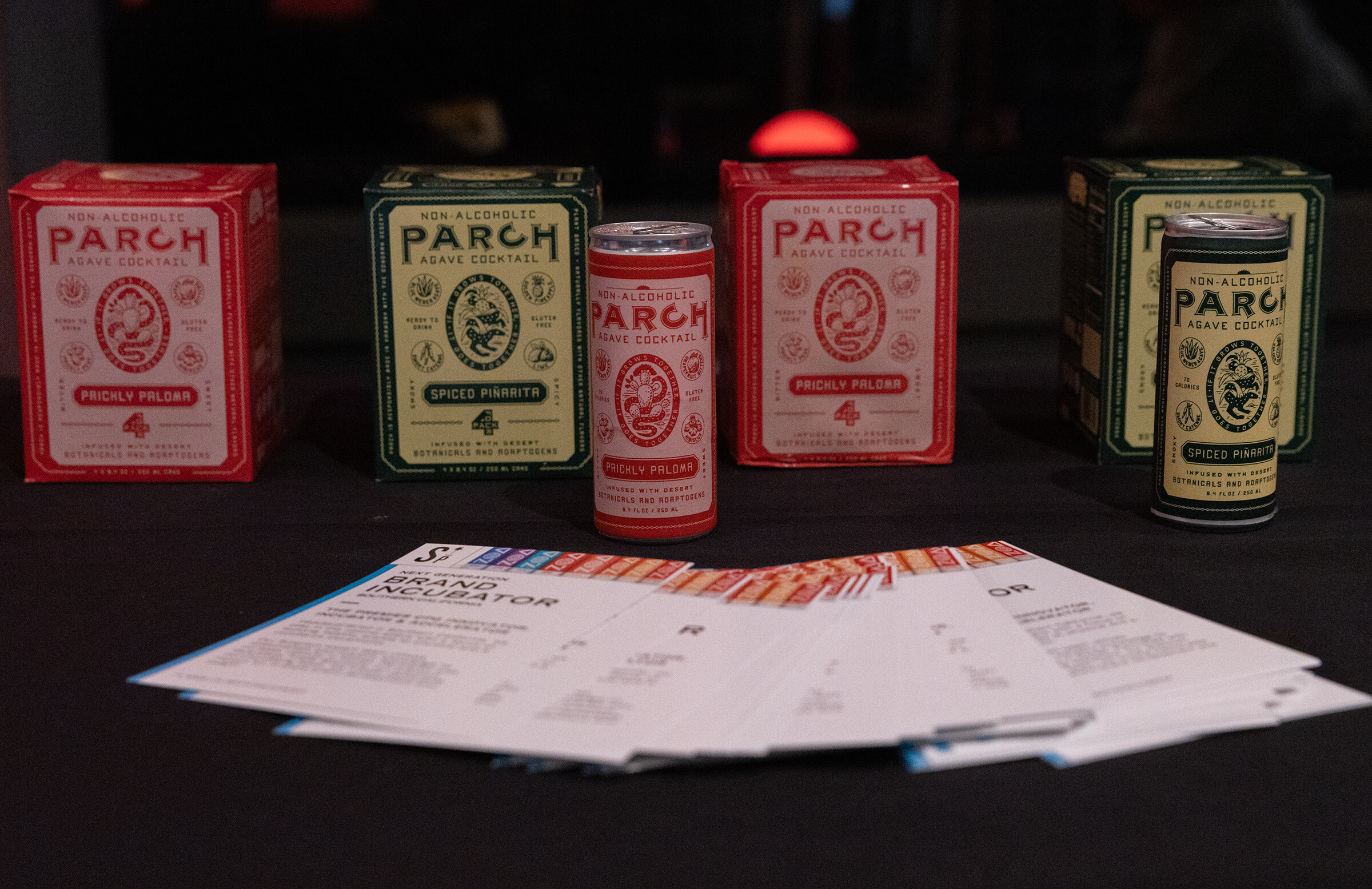 Red and green boxes and cans of Parch non-alcoholic cocktails on a black table with printed brochures.