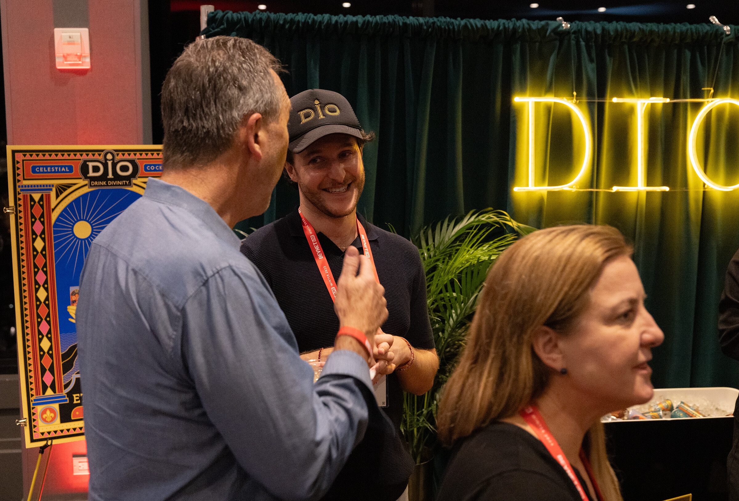 Two men and a woman talk in front of a green curtain with a lit yellow neon sign.