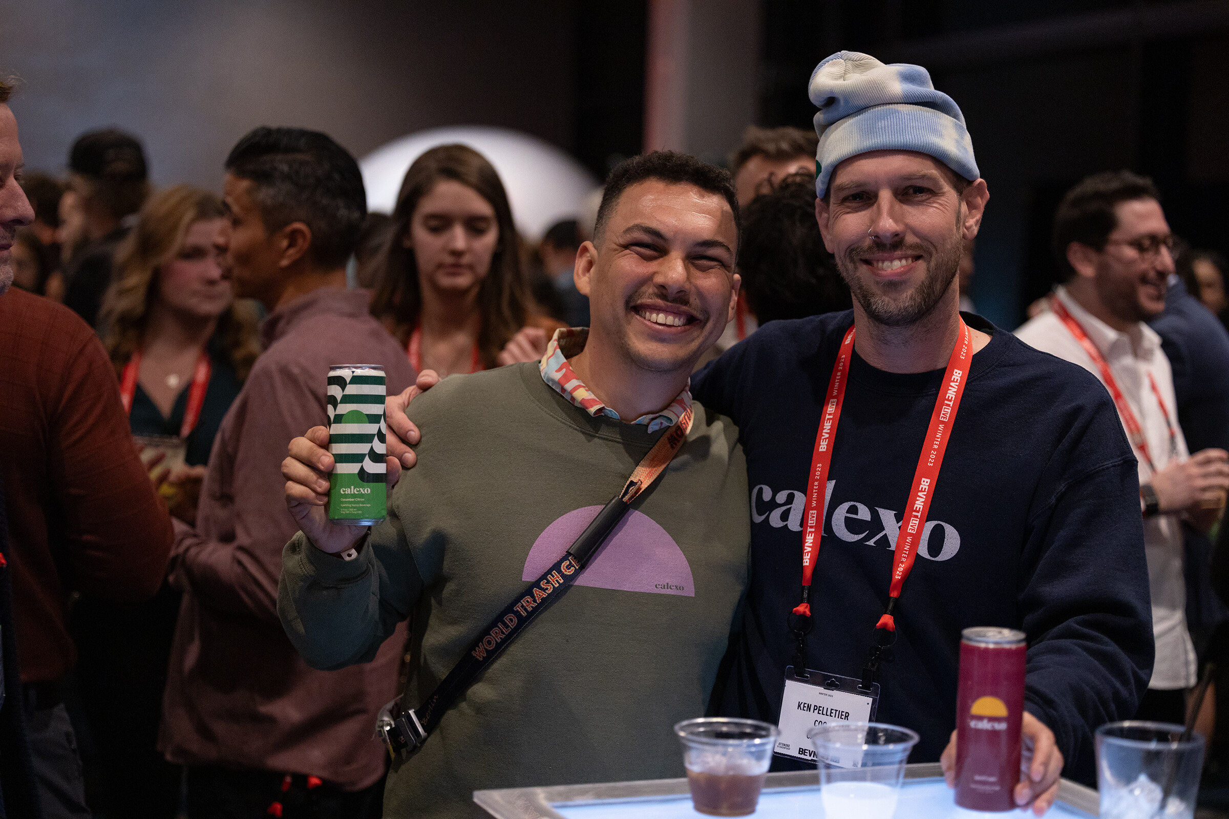 Two men smile and hold cans in front of a crowd; one wears a calexo sweatshirt.