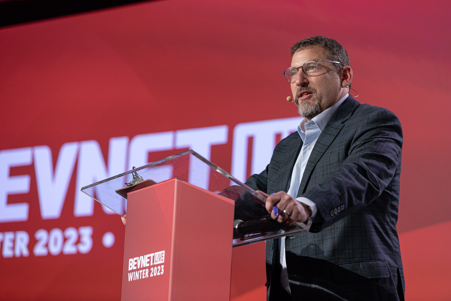 A man in a dark jacket and glasses speaks at a red lectern in front of a BevNET Live screen.