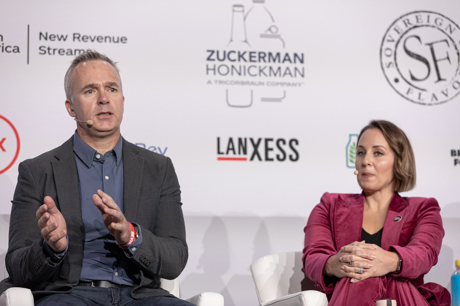 A man in a gray jacket and a woman in a magenta blazer sit on stage in front of a sponsor wall.