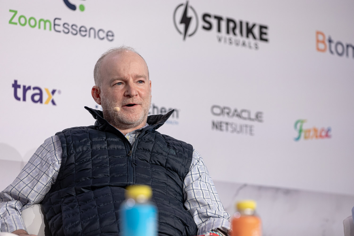 A man in a navy puffer vest and plaid shirt speaks in front of a sponsor wall.