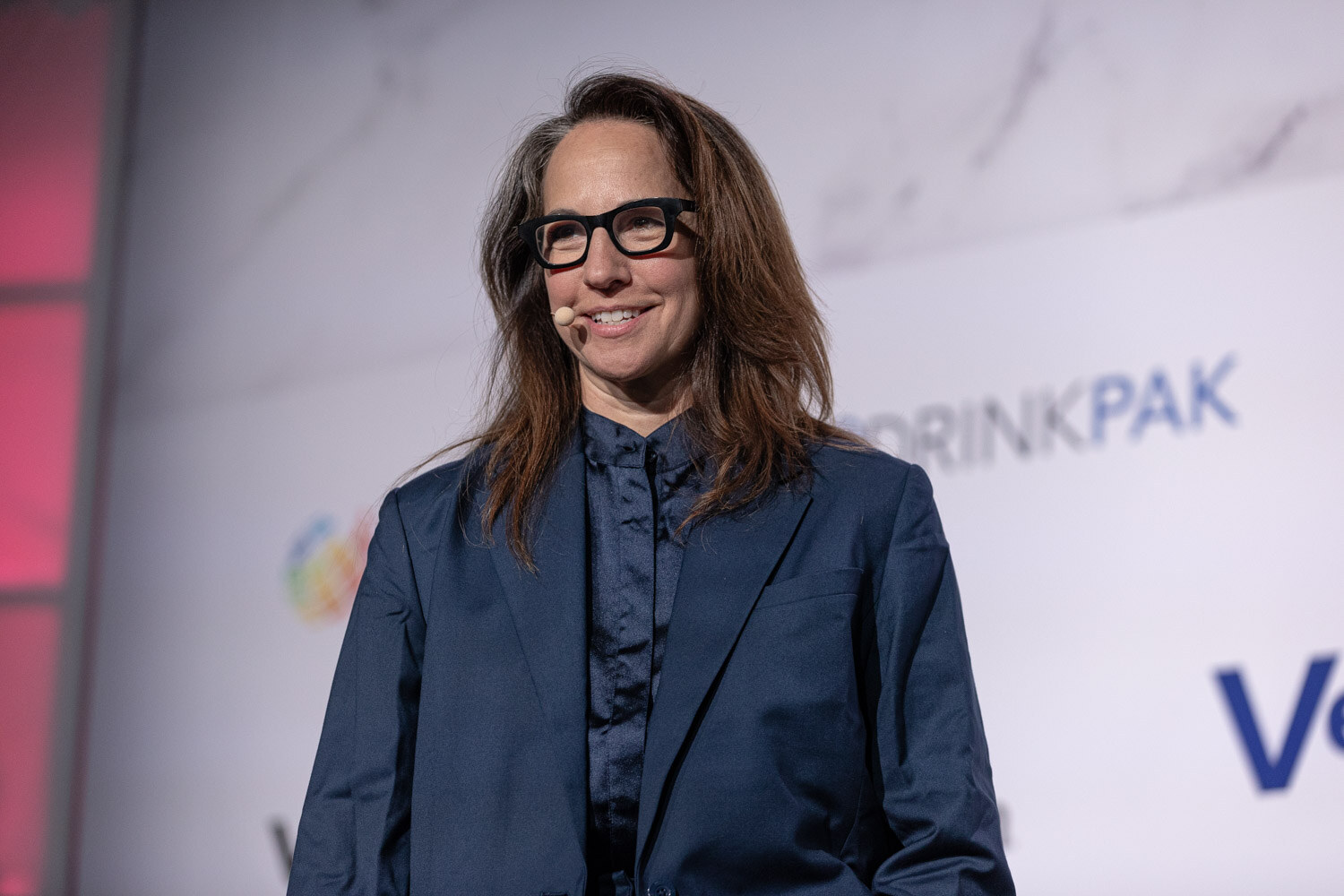 A woman with long hair and black glasses in a navy suit speaks in front of a sponsor wall.