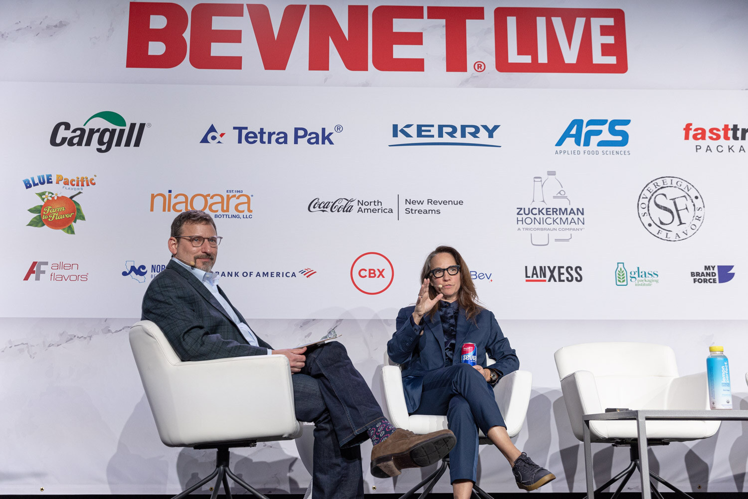 A man in a dark suit and a woman in a navy suit sit in white chairs on stage in front of a BevNET Live sponsor wall.
