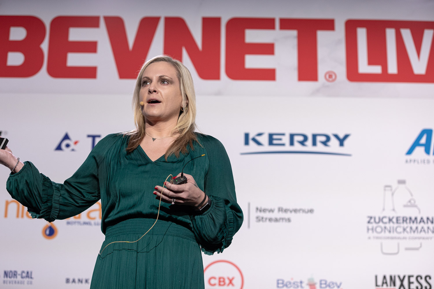A woman in a green dress speaks and gestures in front of a BevNET Live sponsor wall.