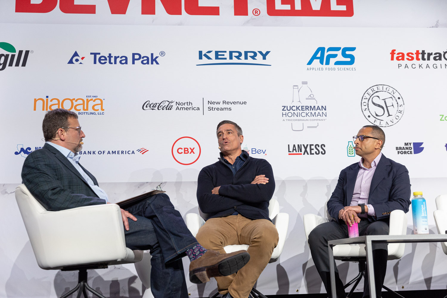 Three men sit in white chairs on a stage in front of a sponsor wall.