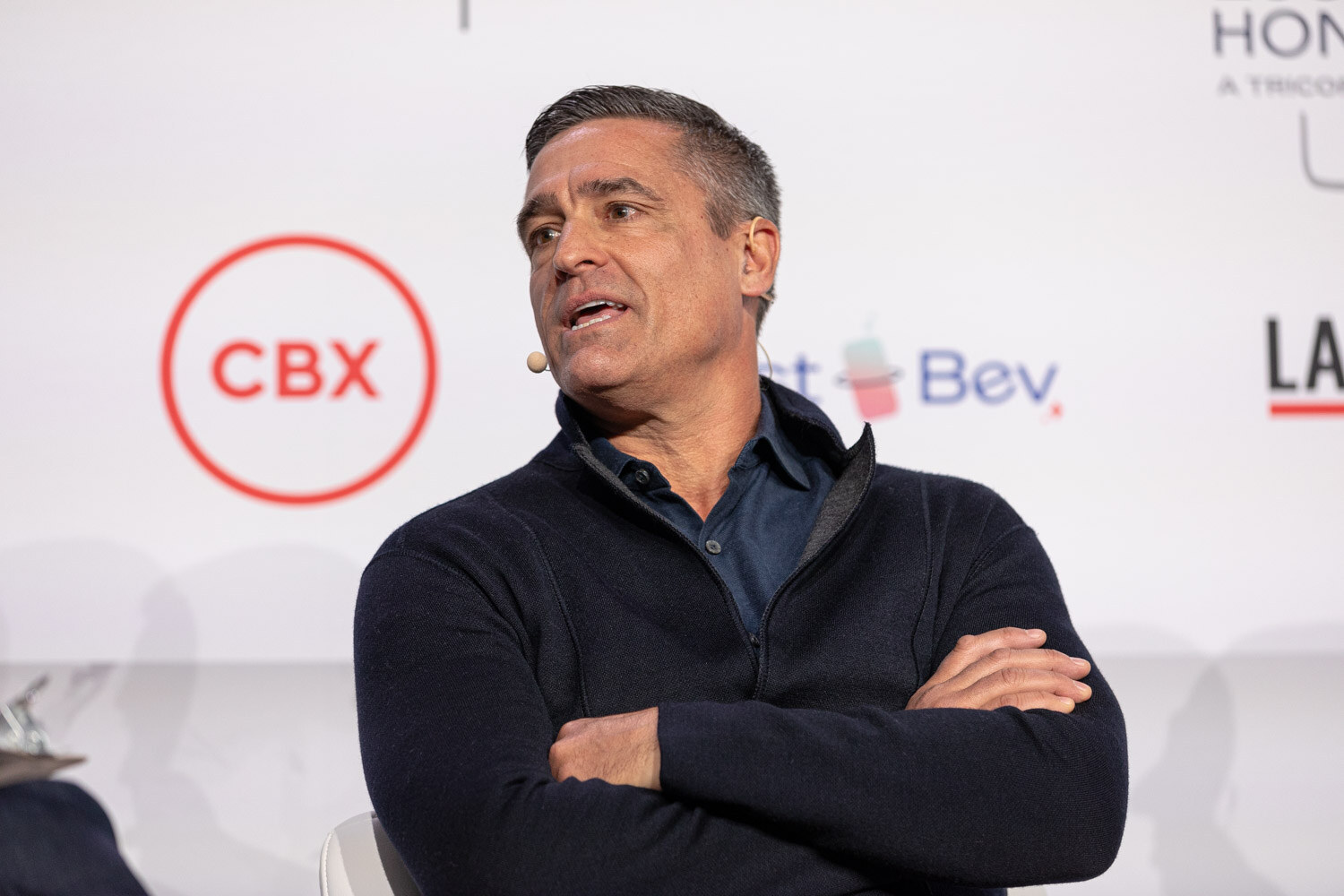 A man with graying hair in a navy sweater sits with arms crossed in front of a CBX sponsor logo.