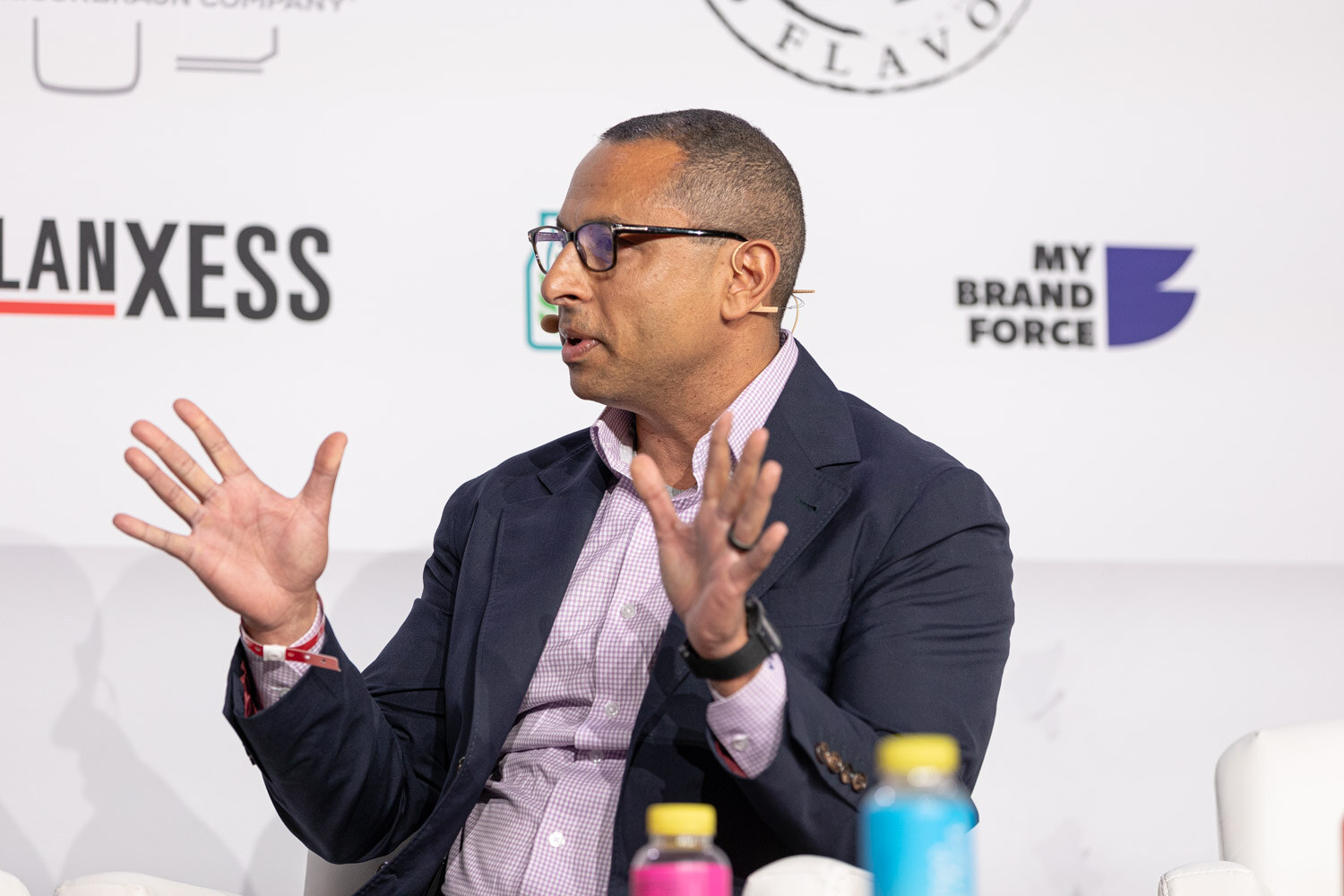 A man in glasses and a navy blazer gestures with both hands while seated on stage.