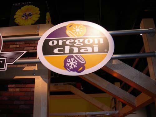 Round Oregon Chai sign mounted on a pipe above a wooden booth frame, with a yellow flower graphic and a brick-patterned wall in the background.