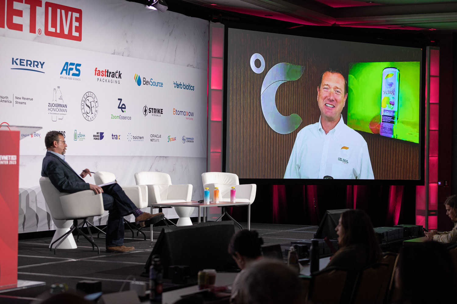 A man seated on stage looks toward a large video screen that shows a person in a white shirt holding a beverage can.