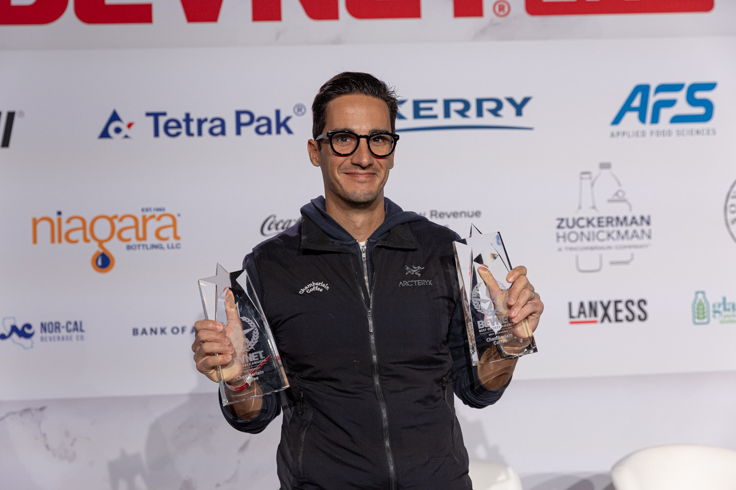 A man in a dark zip jacket holds two clear glass award trophies on stage in front of a sponsor backdrop.