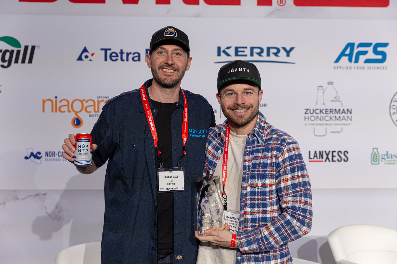 Two men in lanyards stand side by side, one holding a beverage can and the other a glass award trophy.