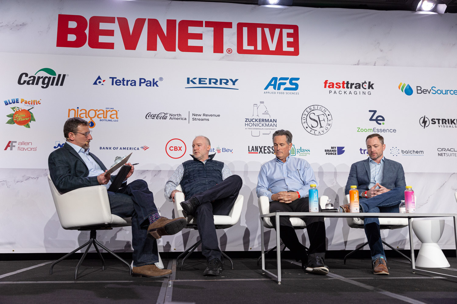 Four people sit in white chairs on a stage under a large BevNET Live sponsor wall.