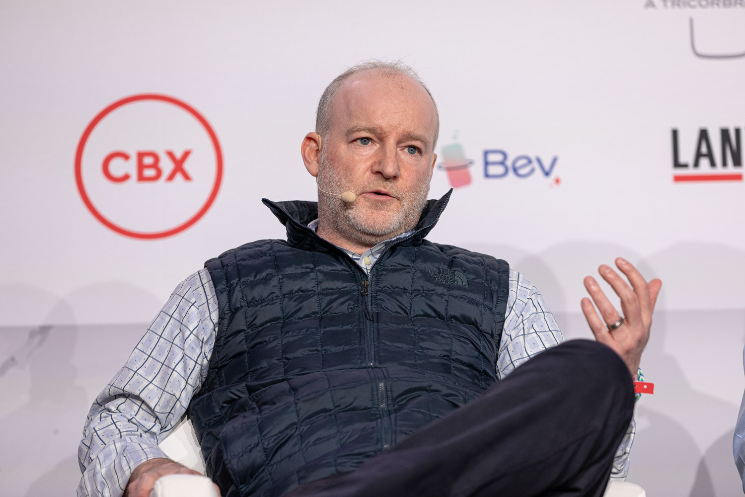 A man with a gray beard in a navy puffer vest gestures while speaking in front of a CBX sponsor logo.