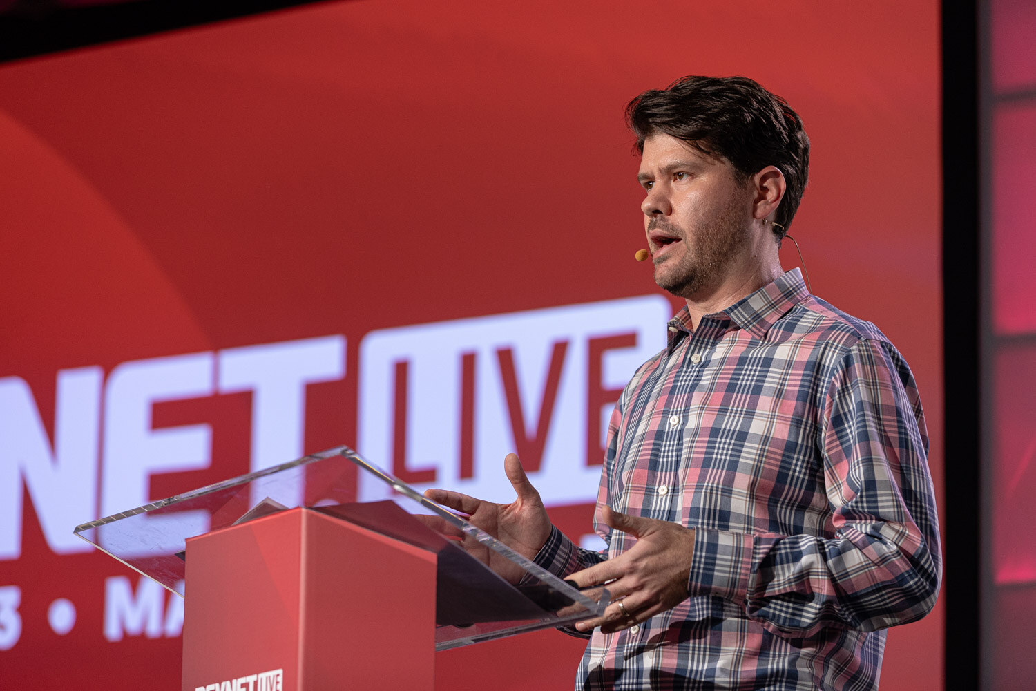A man in a plaid shirt speaks at a red lectern in front of a BevNET Live screen.