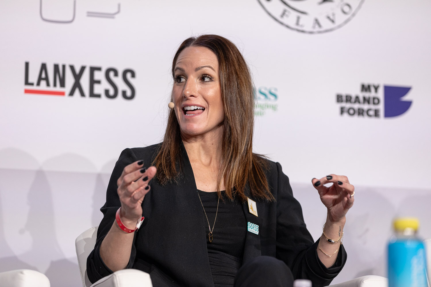 A woman in a black blazer speaks with both hands raised in front of a Lanxess sponsor logo.
