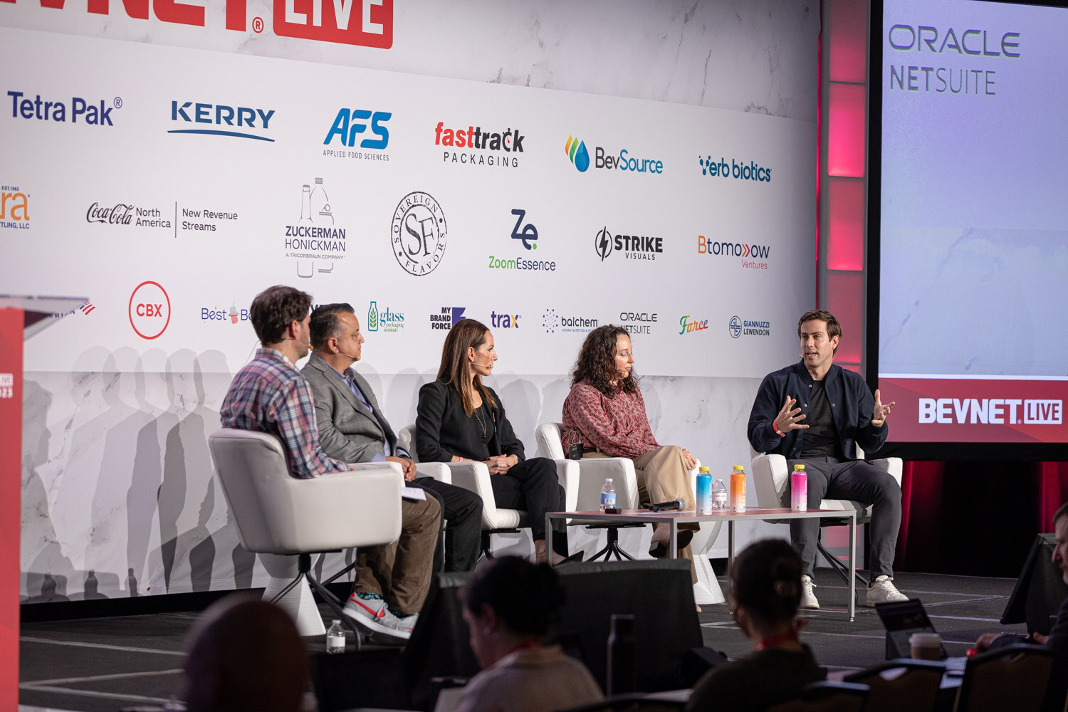 Five people sit in white chairs on a stage in front of a sponsor wall with the audience at the bottom.