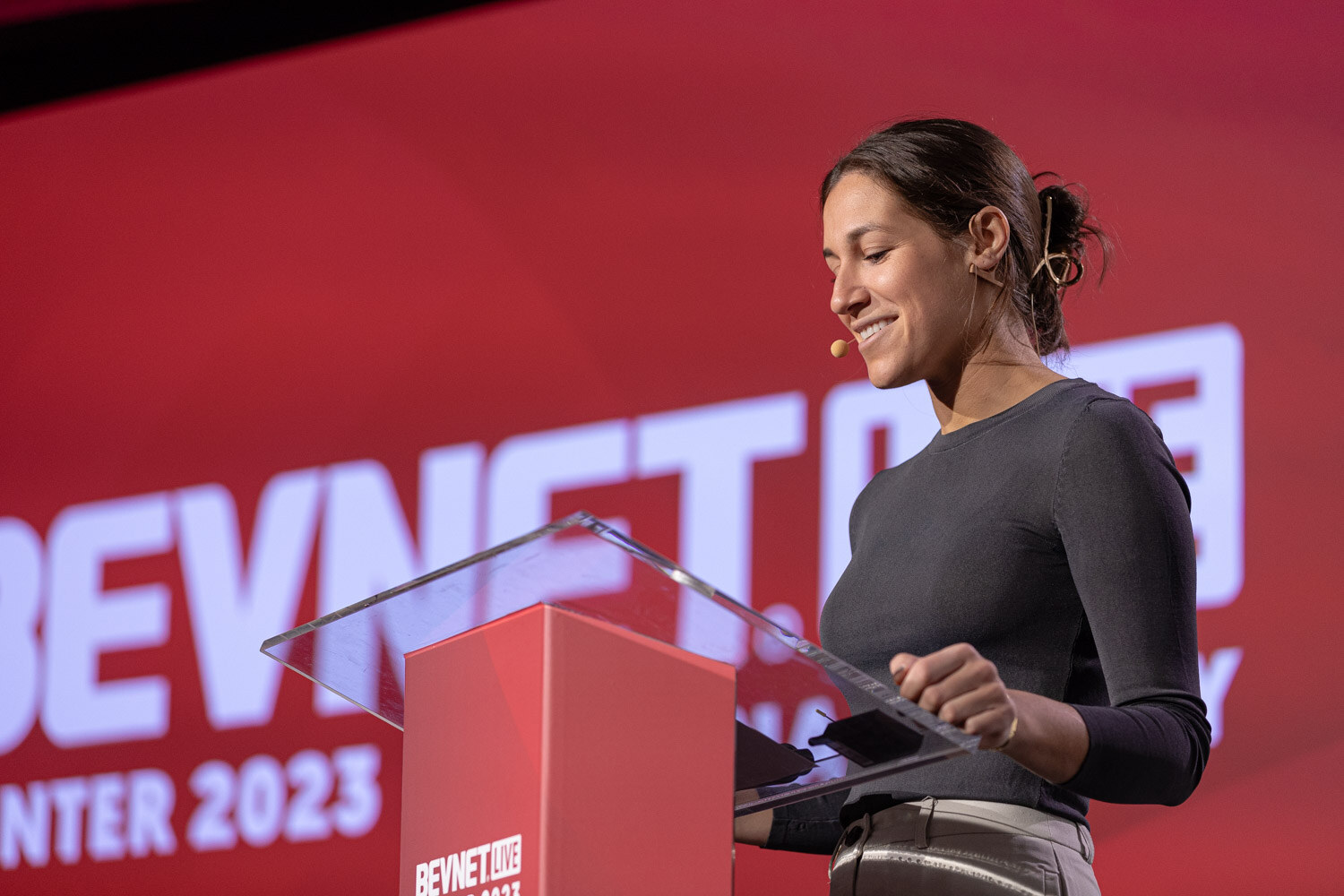 A woman in a gray top looks down at a red lectern in front of a red BevNET Live screen.