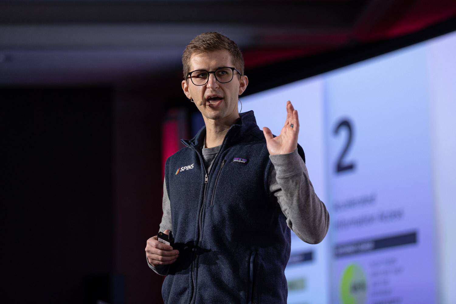 A man in glasses and a SPINS navy vest gestures in front of a slide.