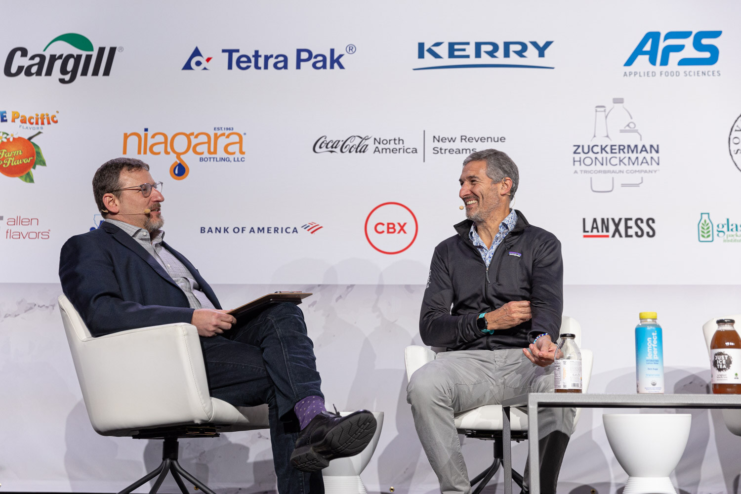Two men sit in white chairs on stage in front of a sponsor wall, talking.