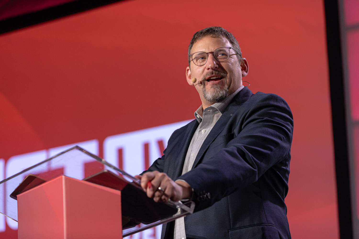 A man with glasses and a goatee in a blue blazer stands at a red lectern against a red screen.