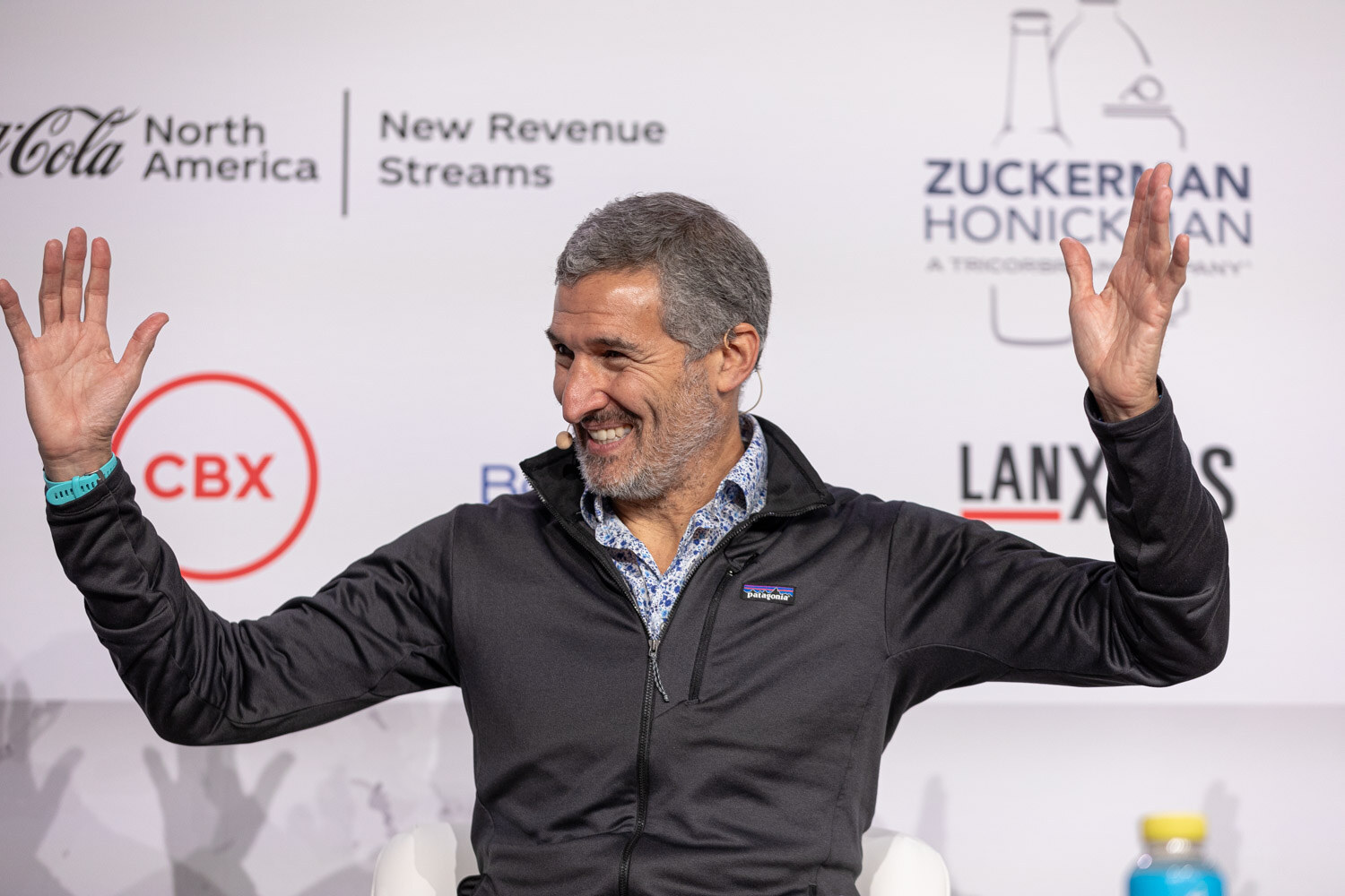 A man in a black jacket smiles with both hands in the air in front of a white backdrop.