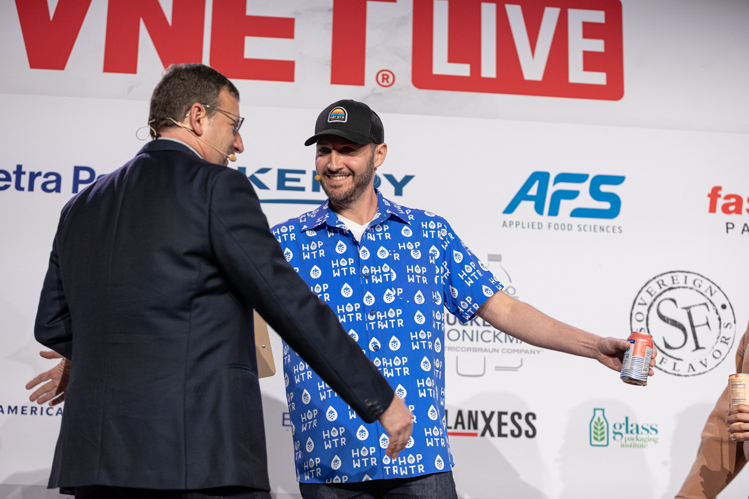 A man in a navy jacket shakes hands with a man in a blue shirt holding a can, in front of a sponsor wall.