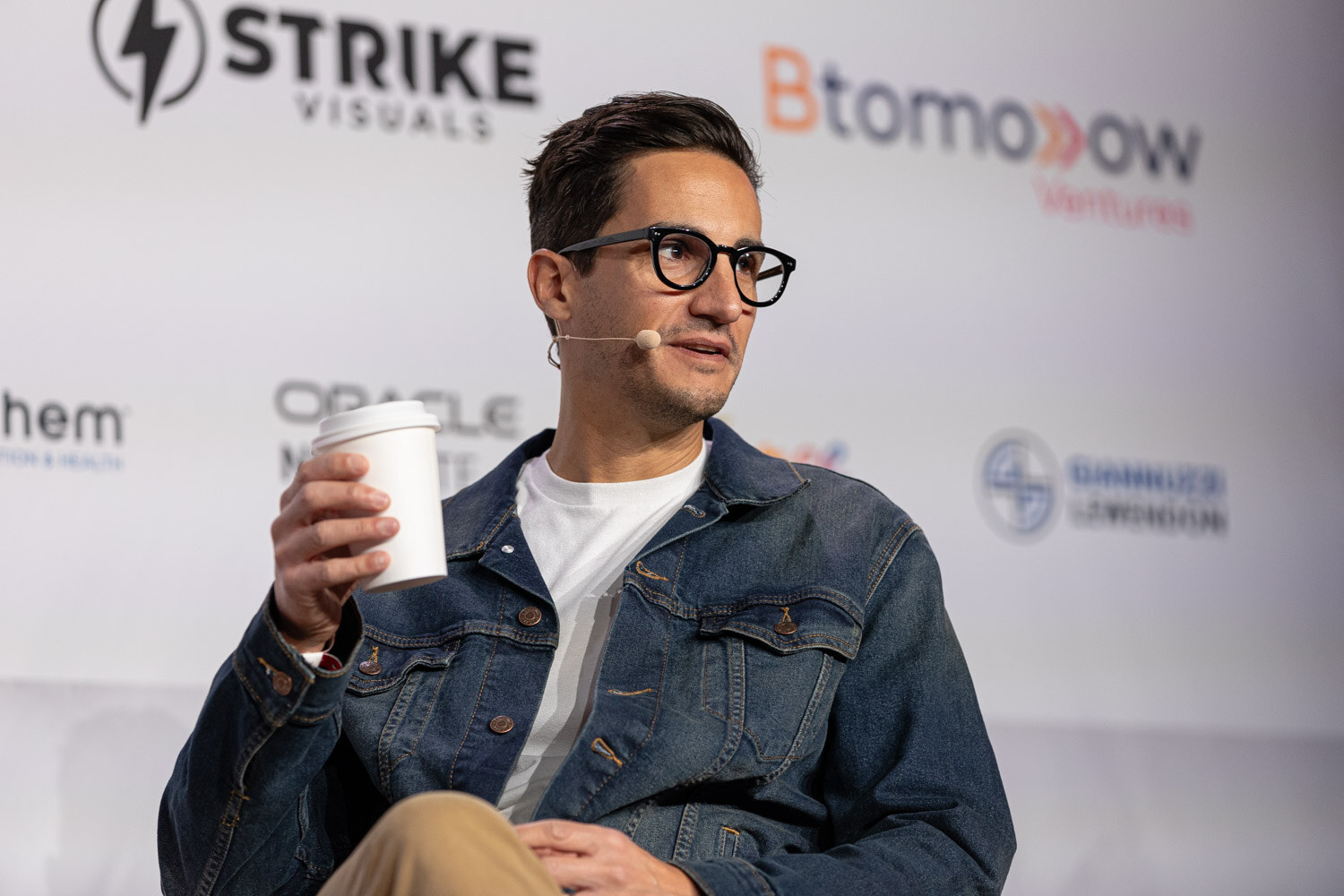 A man in glasses and a denim jacket holds a paper cup and speaks.