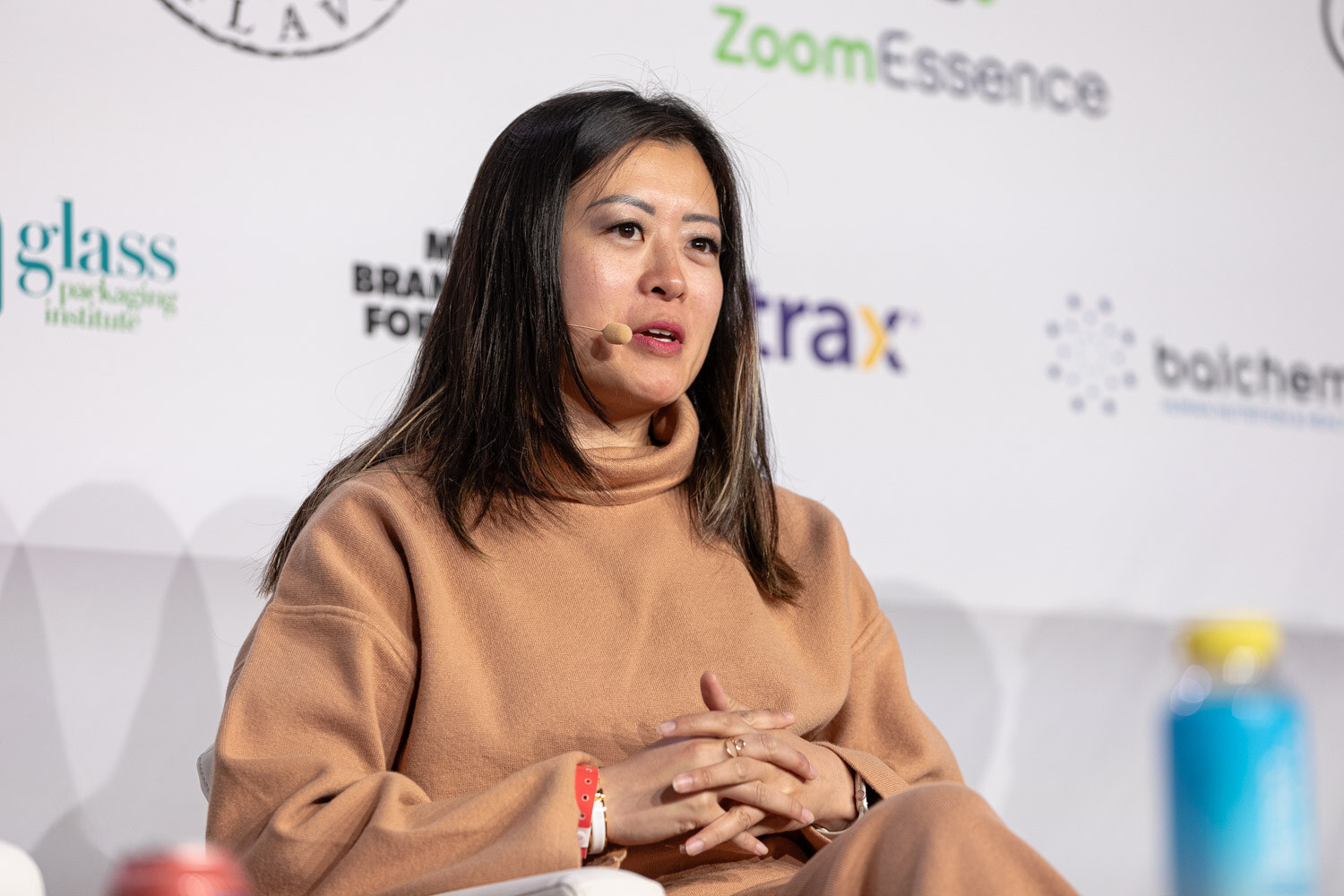 A woman with long dark hair in a tan sweater sits in front of a ZoomEssence sponsor logo.