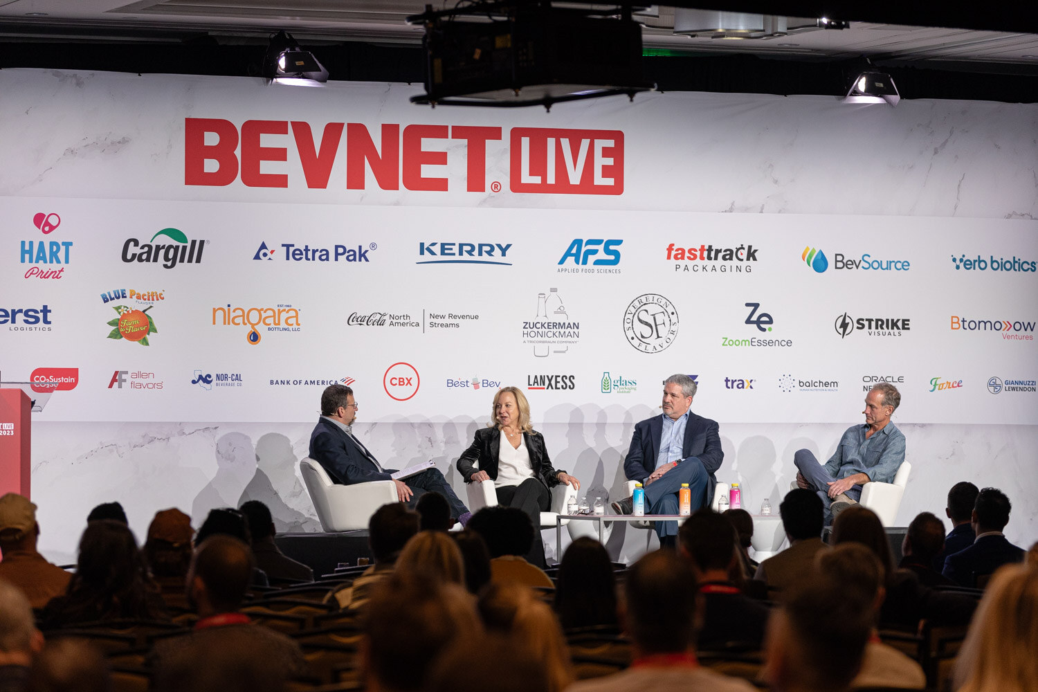 Four people sit in white chairs on a stage under a sponsor wall.