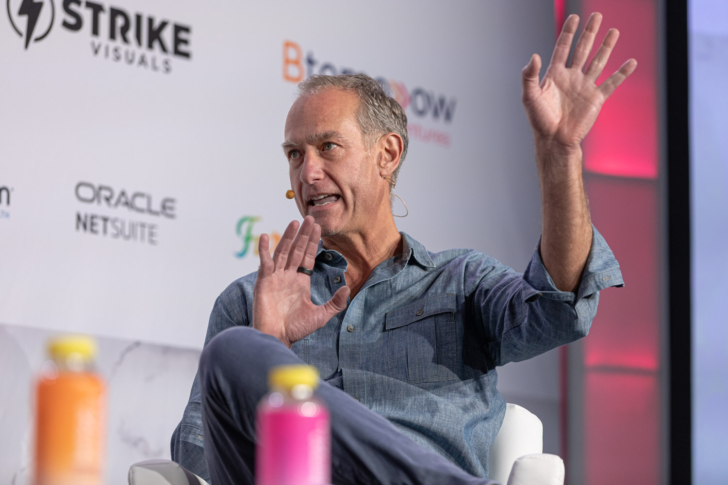 A man in a blue shirt raises both hands while seated on stage in front of Strike Visuals and Oracle NetSuite logos.