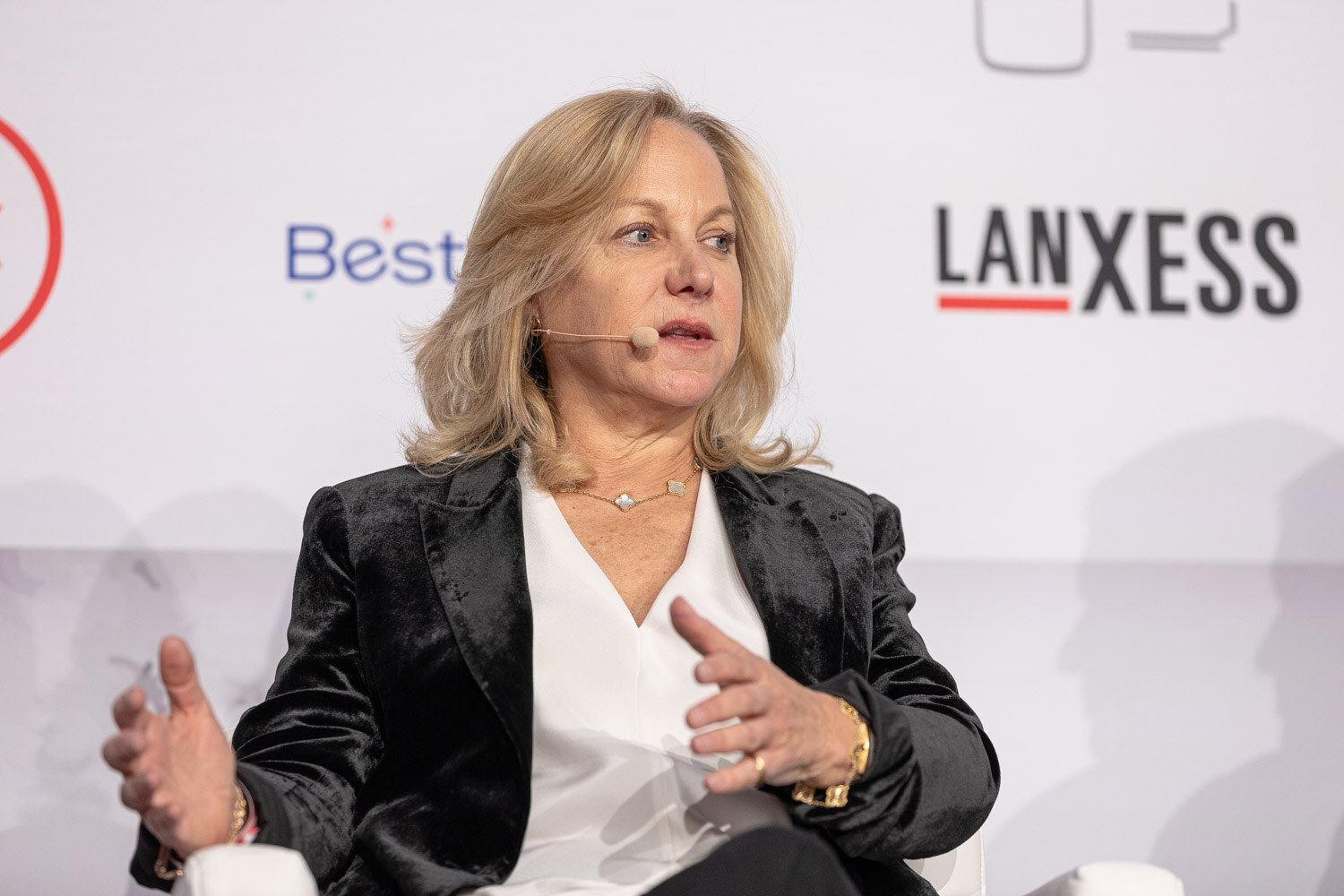 A woman with blond hair in a black blazer and white blouse speaks while seated.