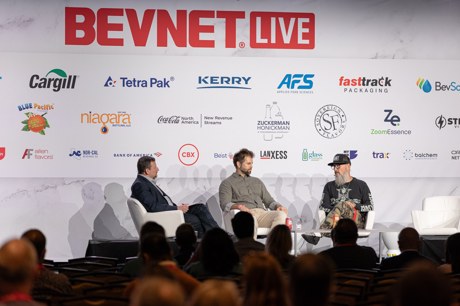 Three people sit on a stage in front of a BevNET Live sponsor wall with an audience in the foreground.