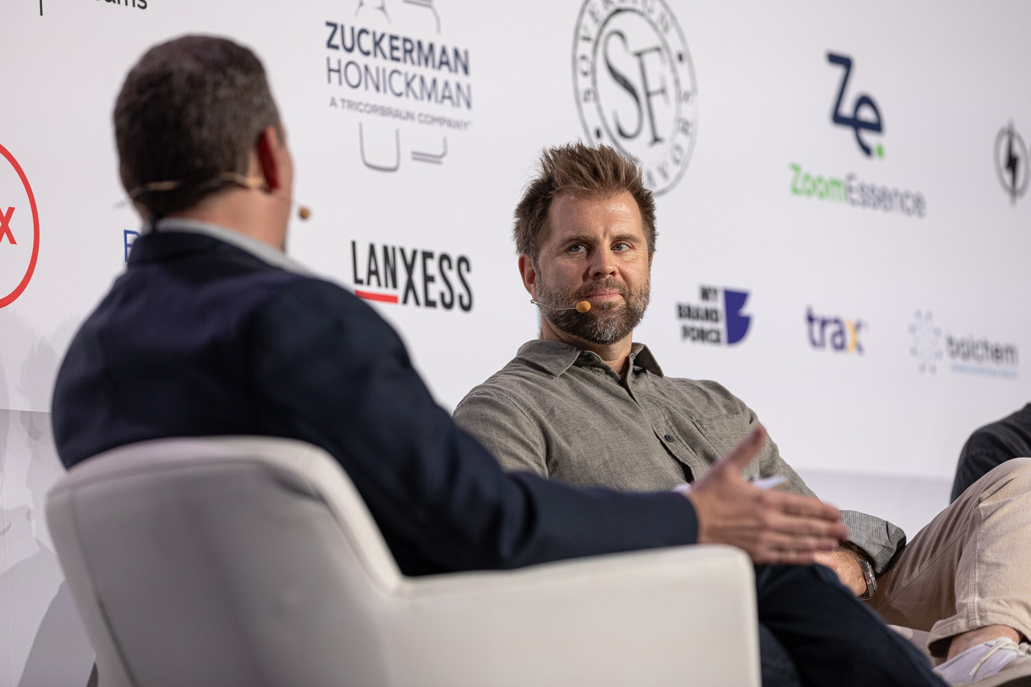 The back of a man's head in the foreground and a man in a gray shirt seated on stage, talking.