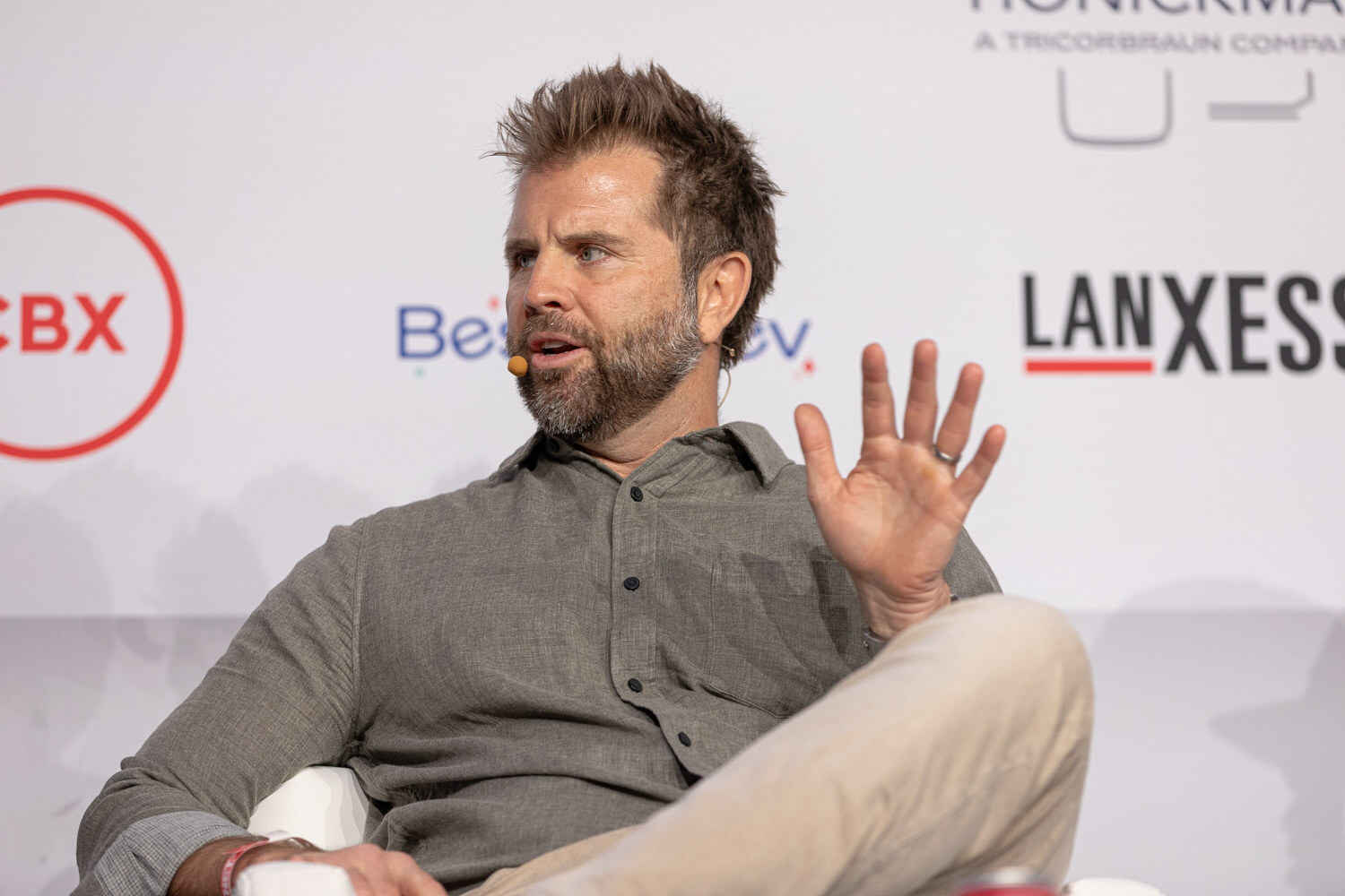 A bearded man in a gray shirt raises one hand while talking in front of a CBX logo.