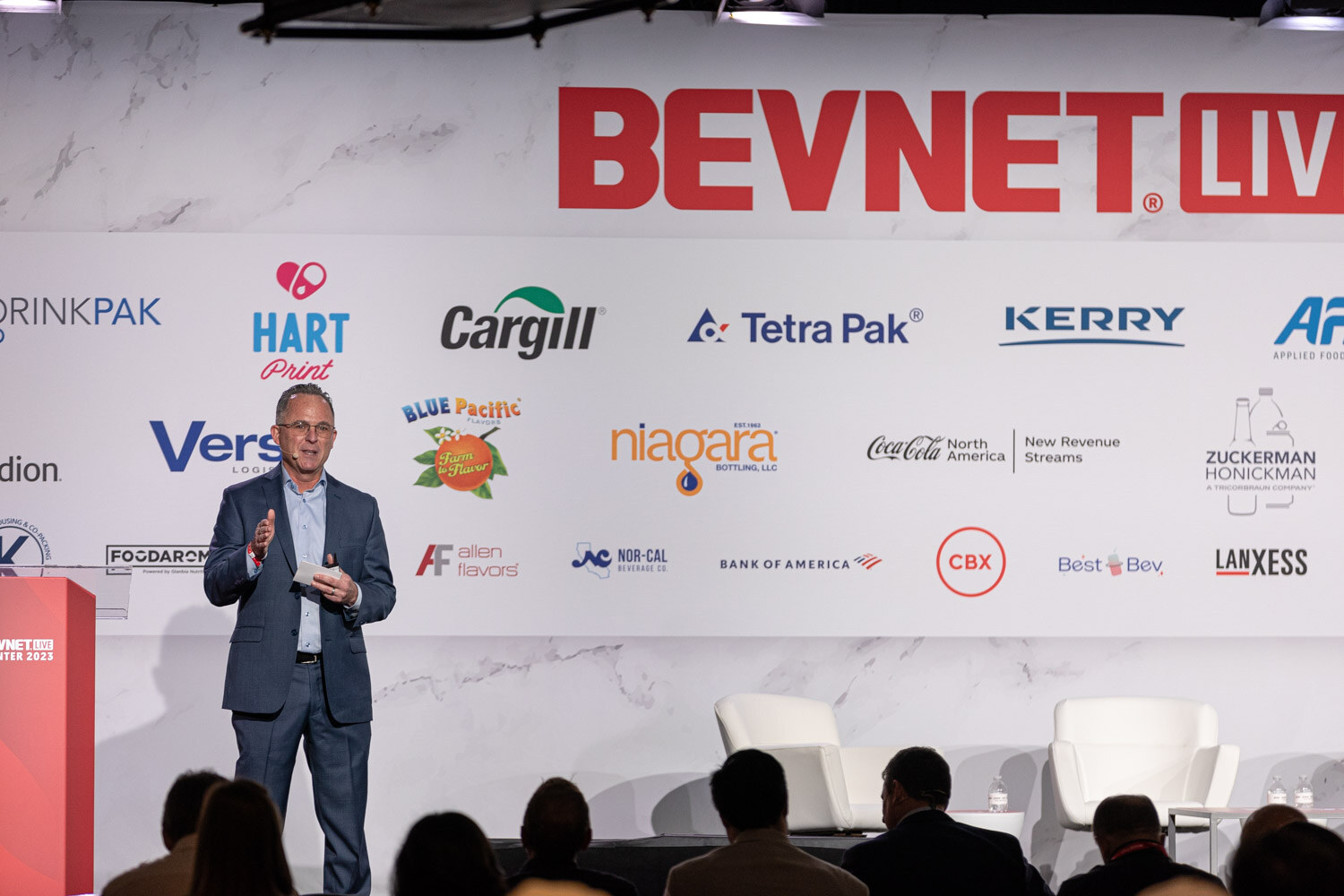 A man in a gray blazer speaks at the mainstage in front of a red sponsor wall.