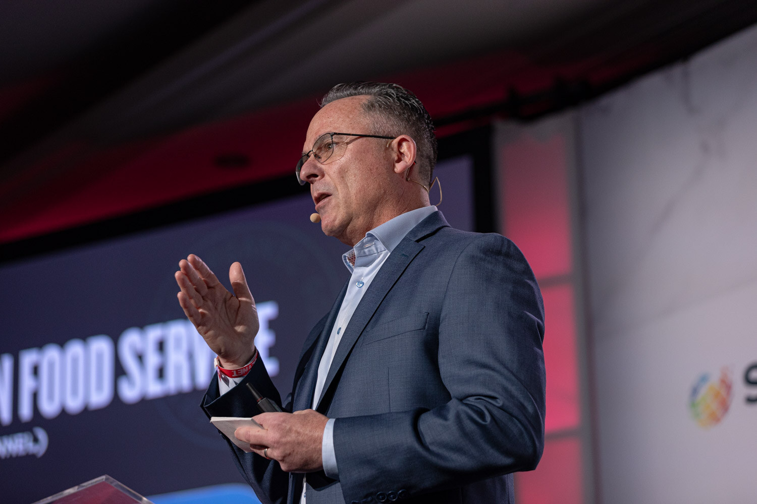 A man in glasses and a gray blazer speaks in front of a slide about food service.