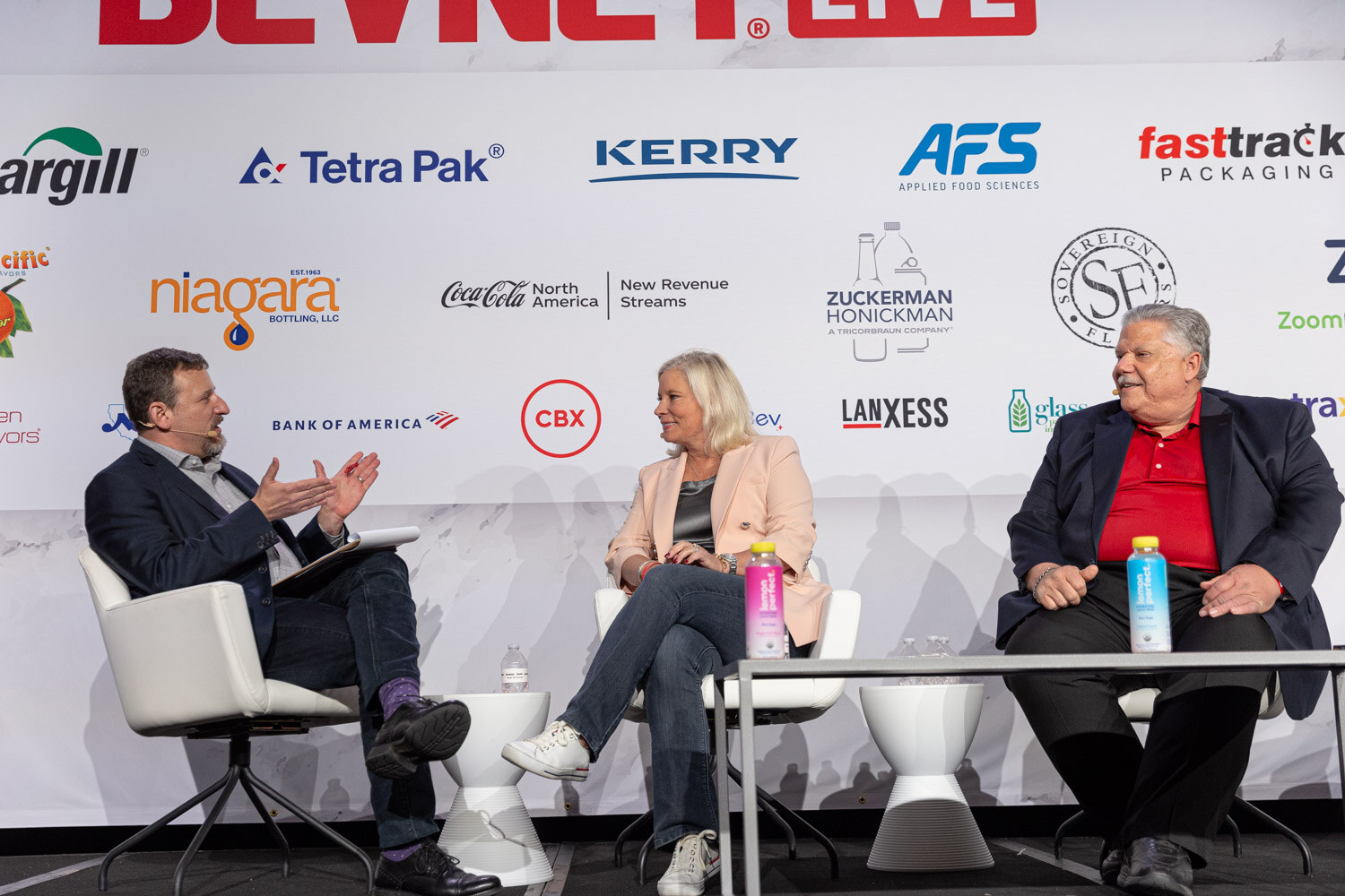 Three people sit in white chairs on a stage in front of a BevNET Live sponsor wall.