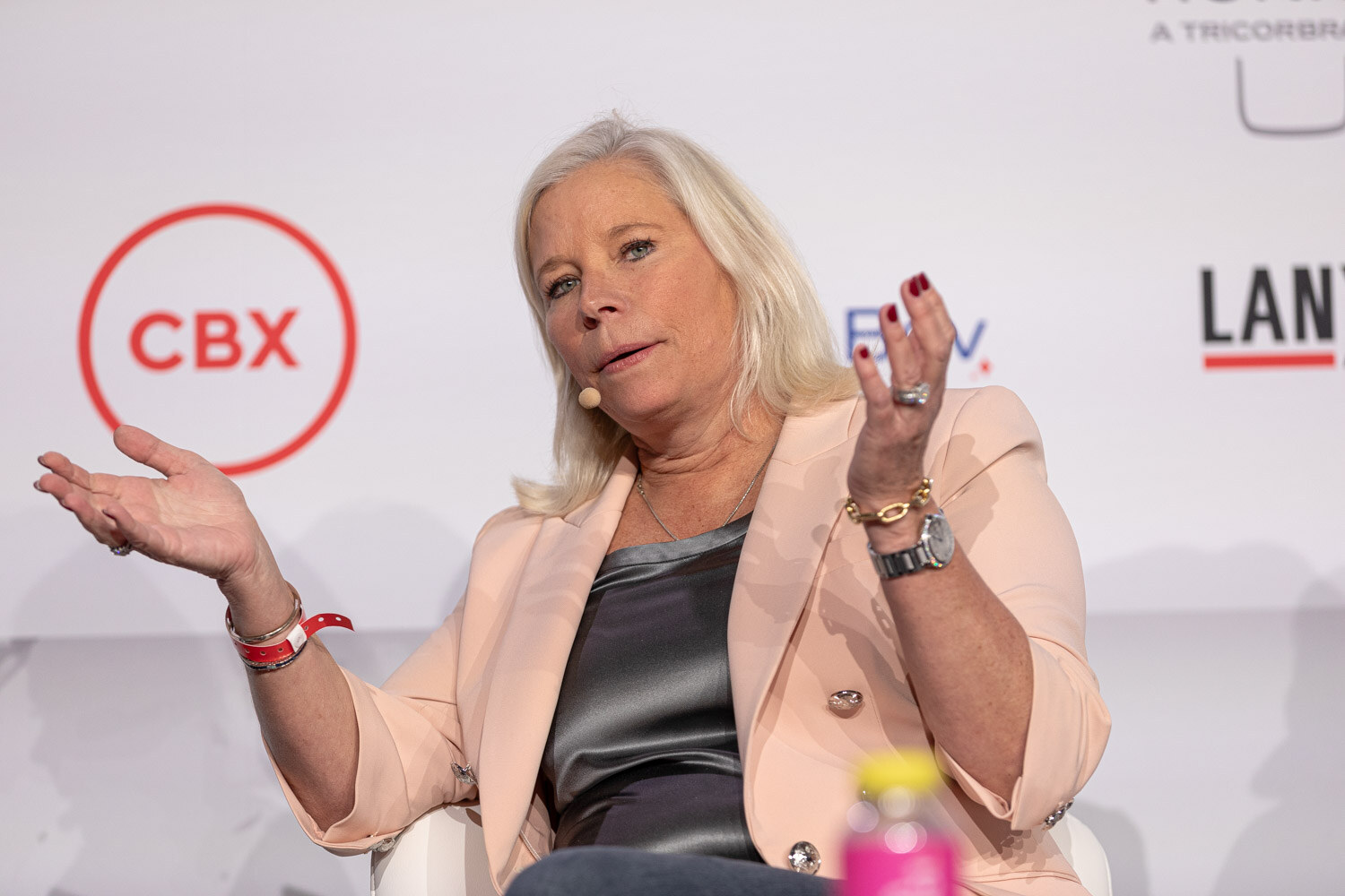 A woman with blond hair in a peach blazer speaks with open hands in front of a CBX sponsor logo.
