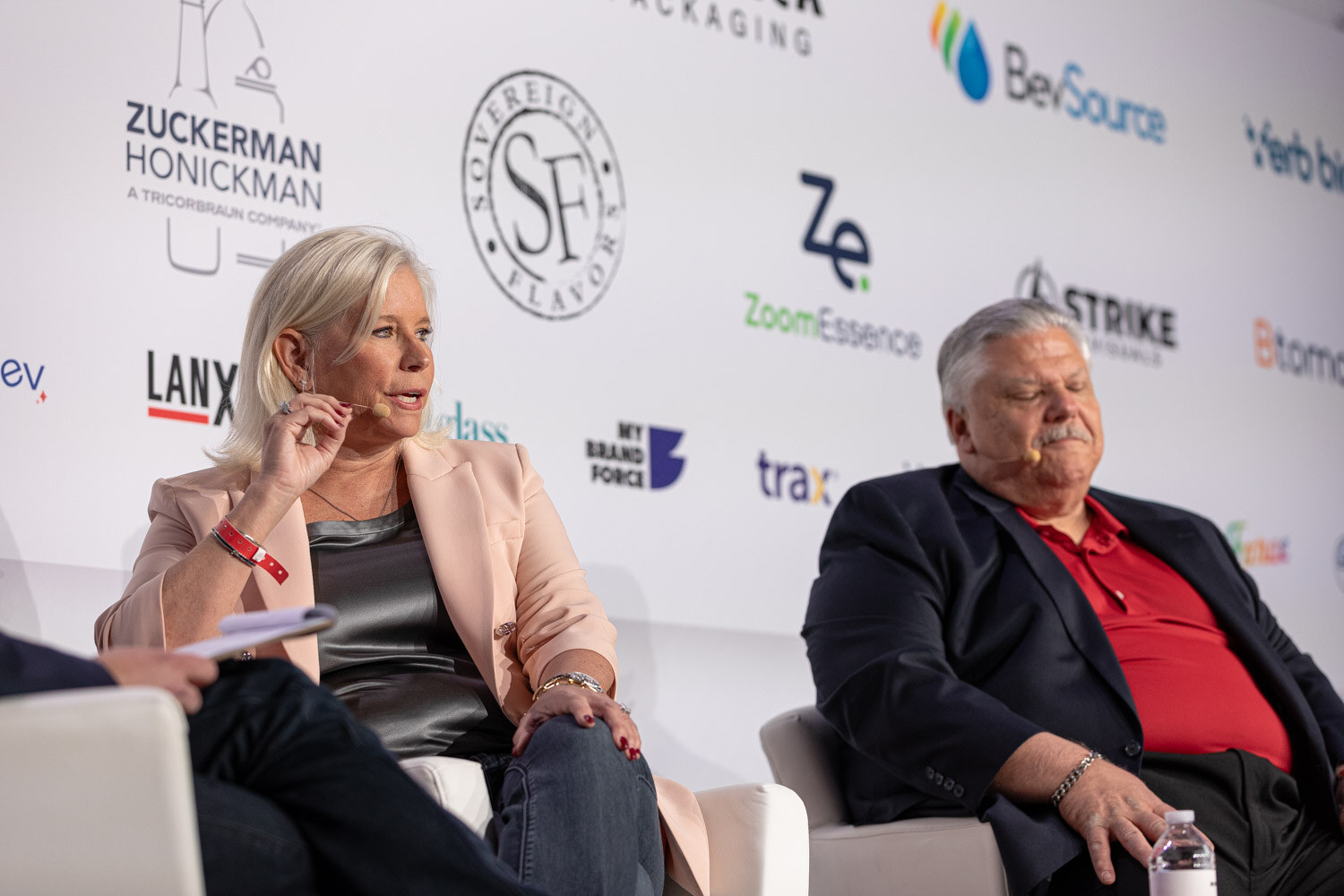 A woman in a peach blazer and a man in a red polo seated on stage beside the back of a moderator's chair.