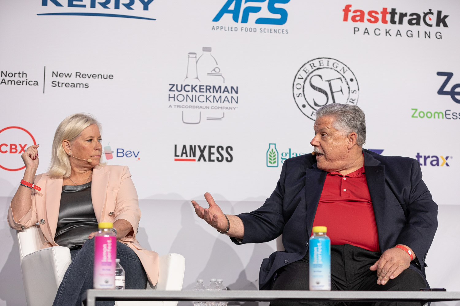 A woman in a peach blazer and a man in a red polo and navy blazer talk on stage with a sponsor backdrop.