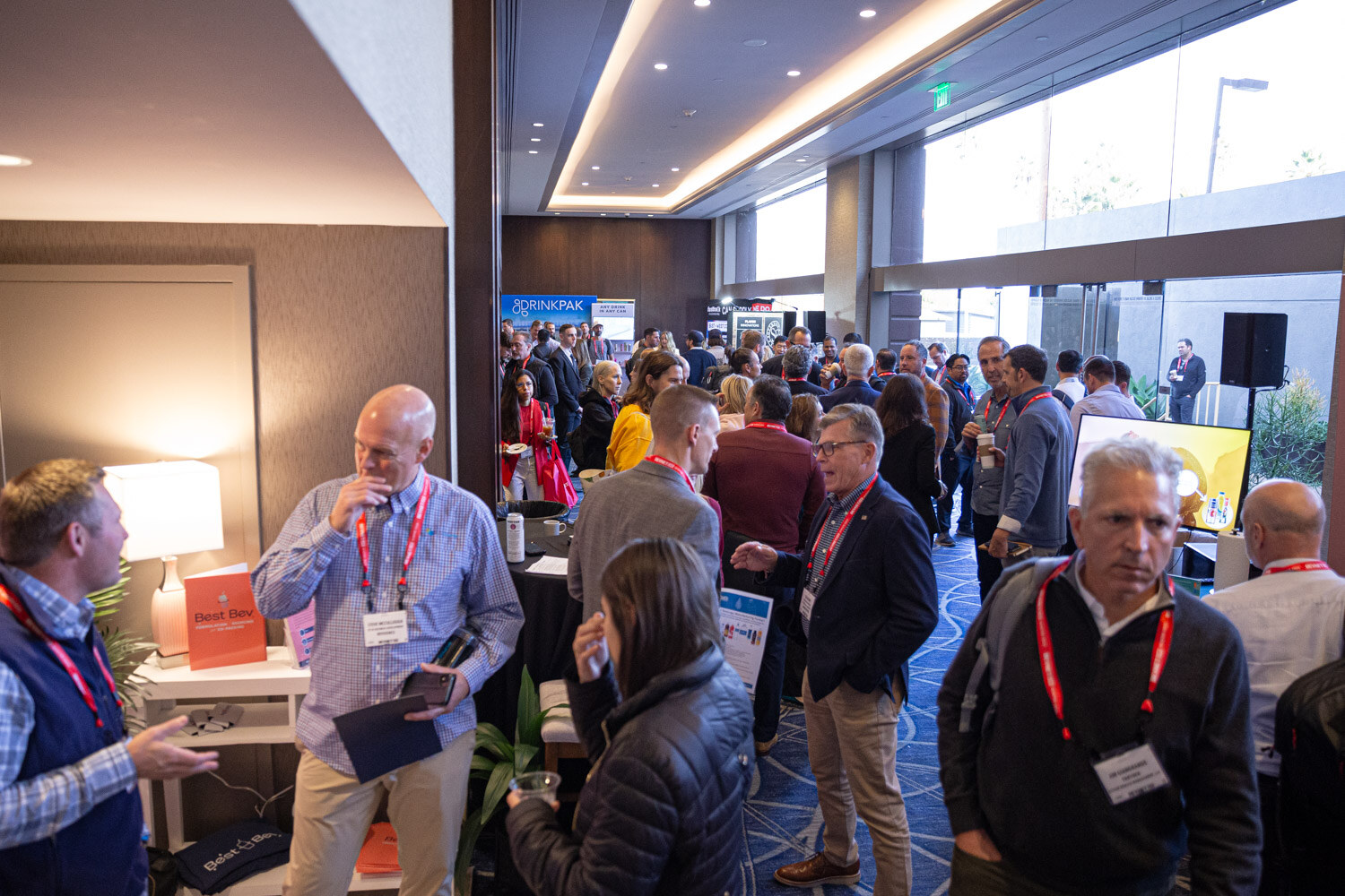 Attendees in lanyards stand and talk near exhibitor tables in a hallway with large windows.