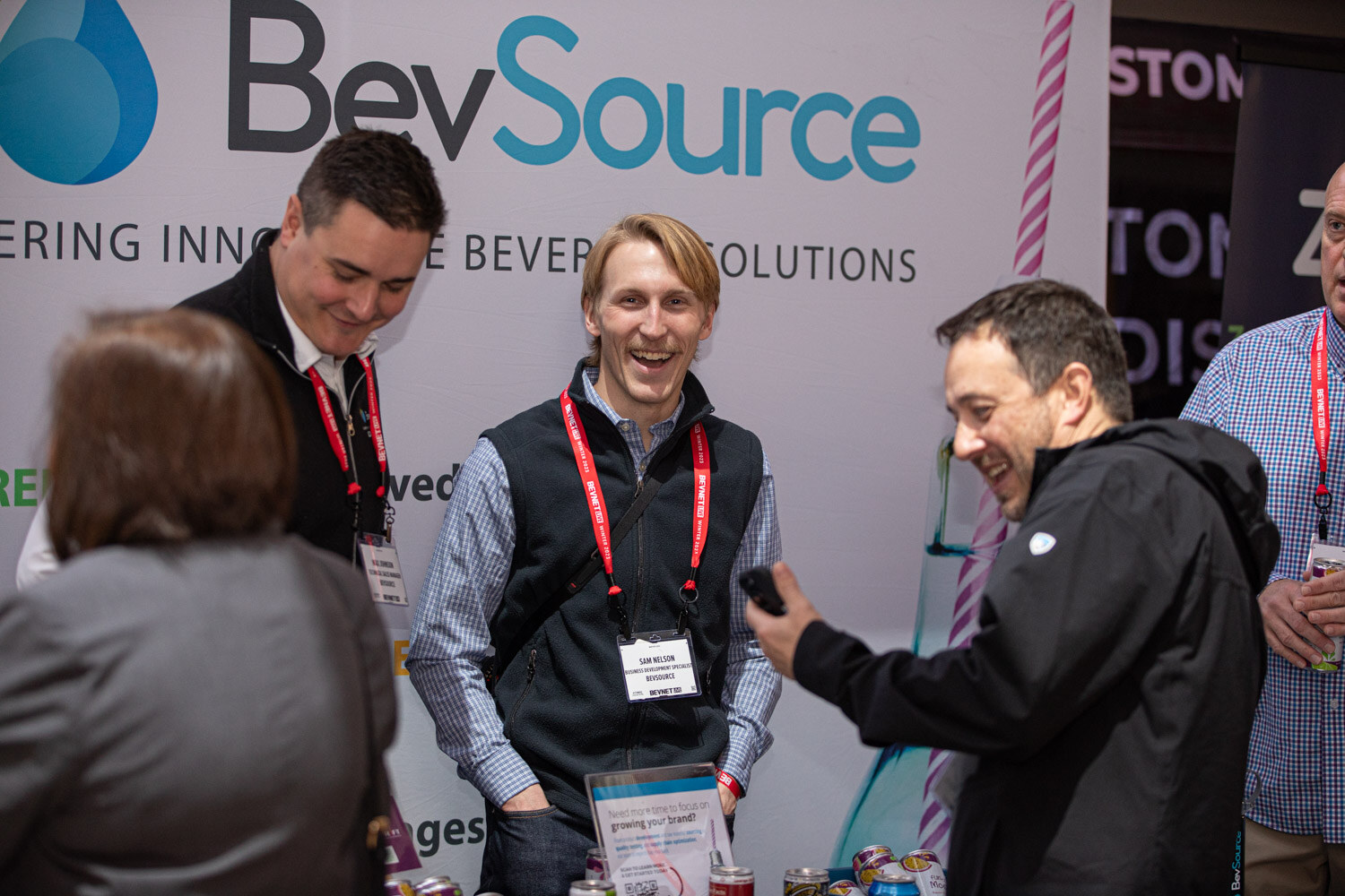 Three men laugh in front of a BevSource banner while other attendees stand around them.