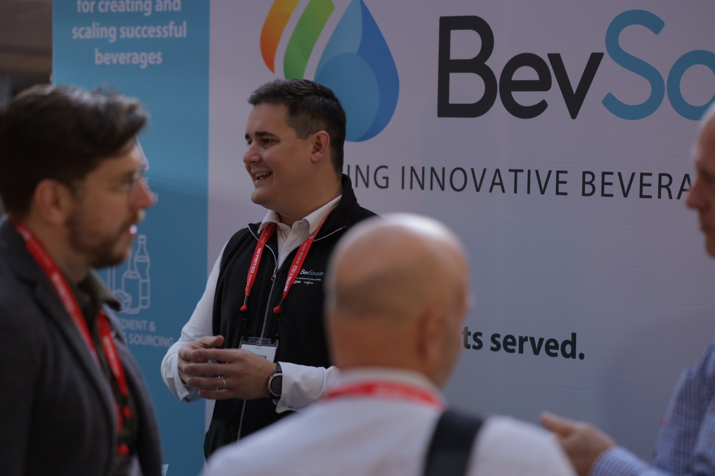 A man in a black vest smiles while talking with two attendees in front of a BevSource banner.