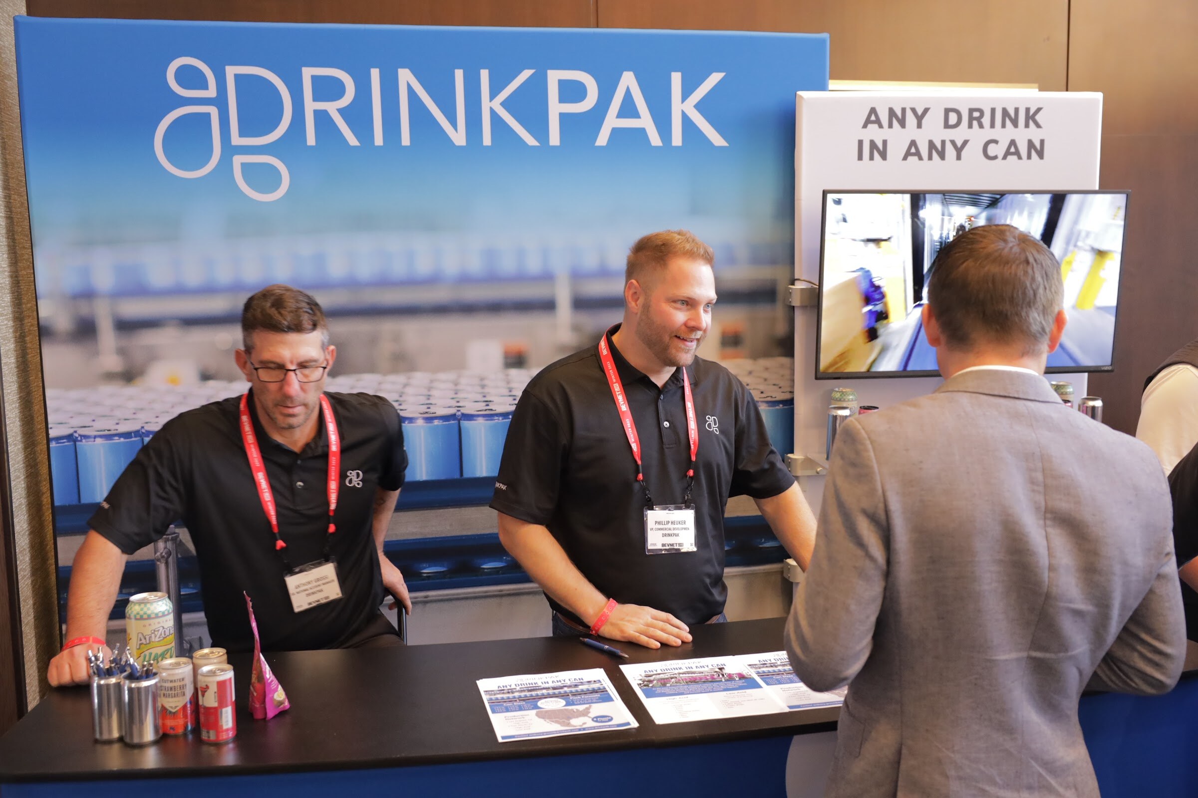 Two men in black shirts stand behind a table with cans and brochures in front of a DrinkPak banner.