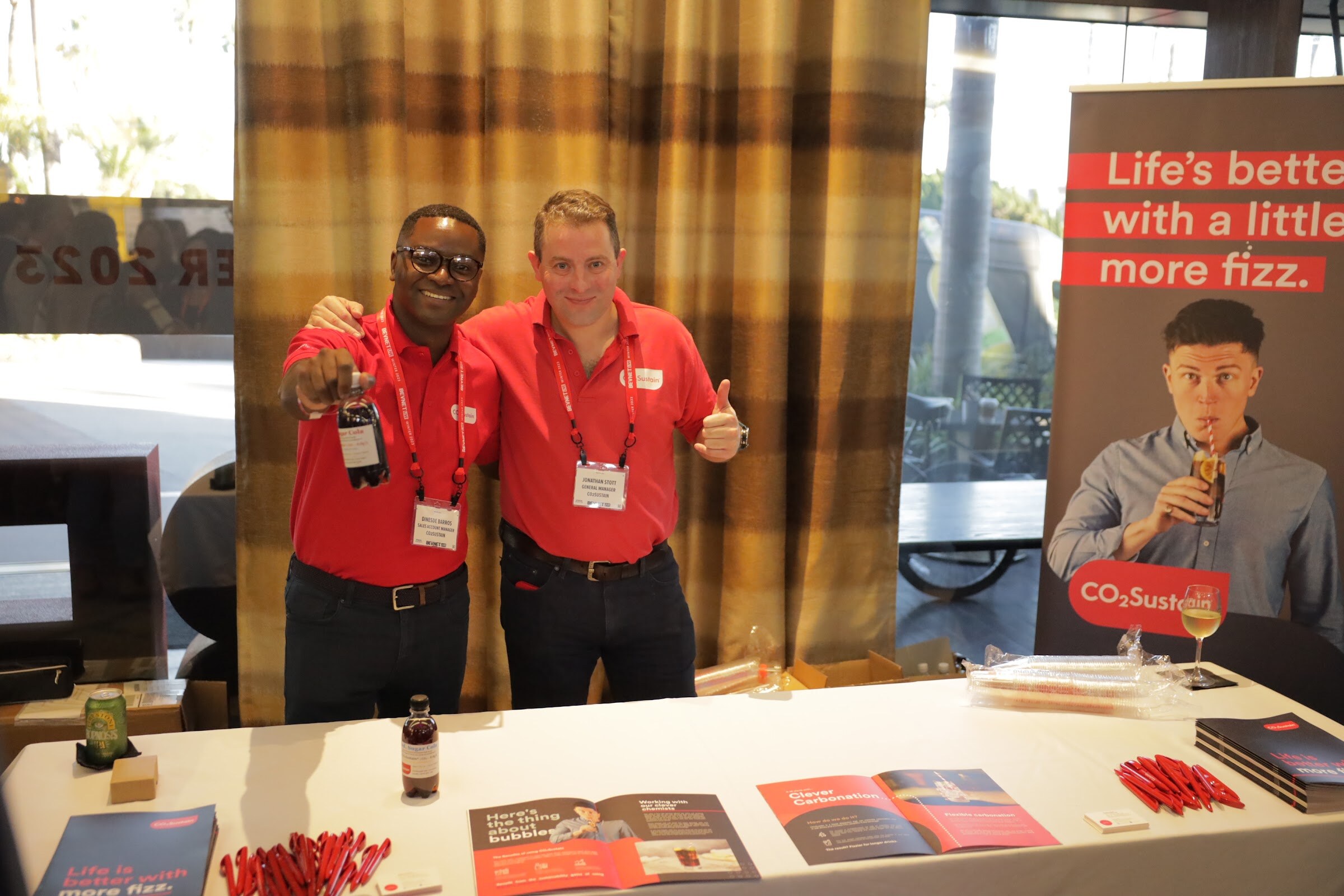 Two men in red polo shirts hold bottles behind a table with brochures and a CO2Sustain banner.