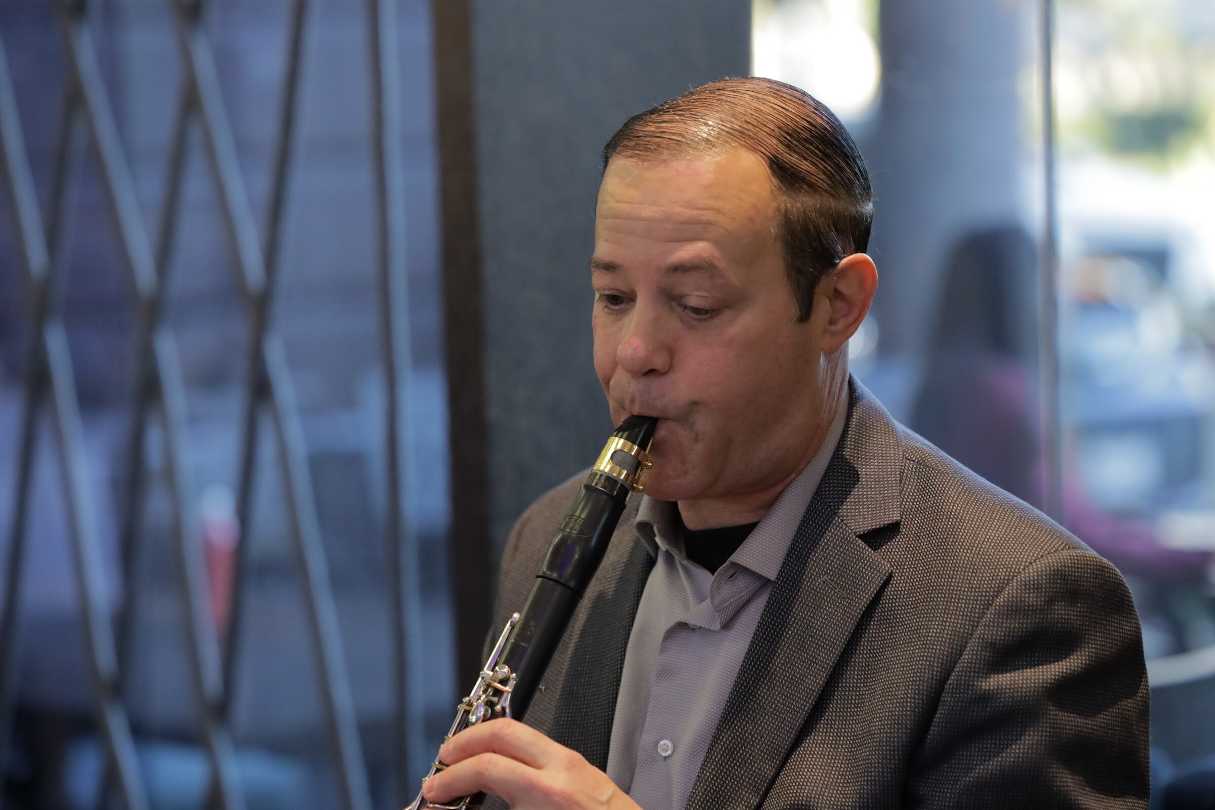 A man in a gray blazer plays a clarinet, with windows and a dark gate behind him.
