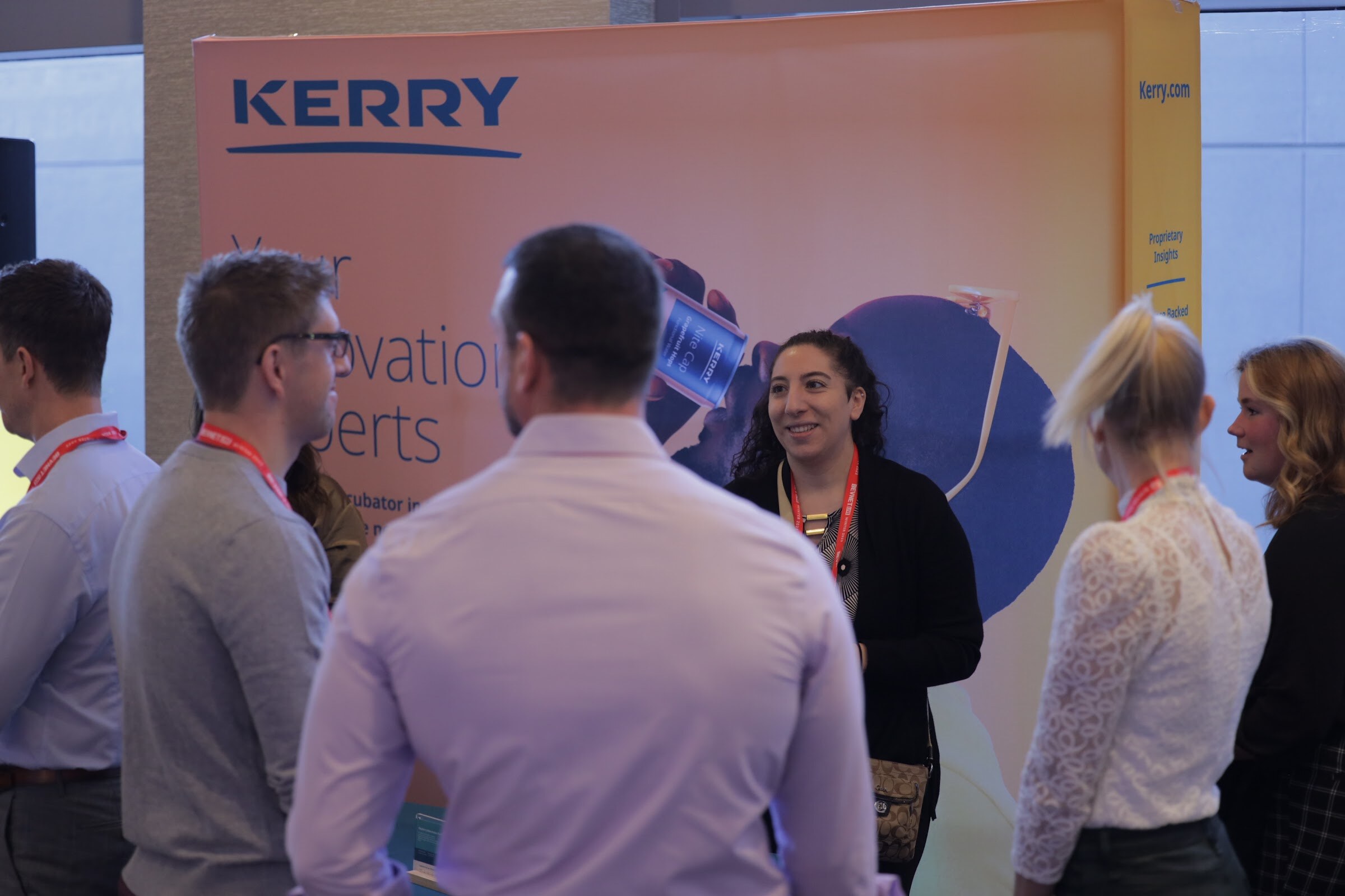 Attendees stand in front of a large orange Kerry banner talking in small groups.